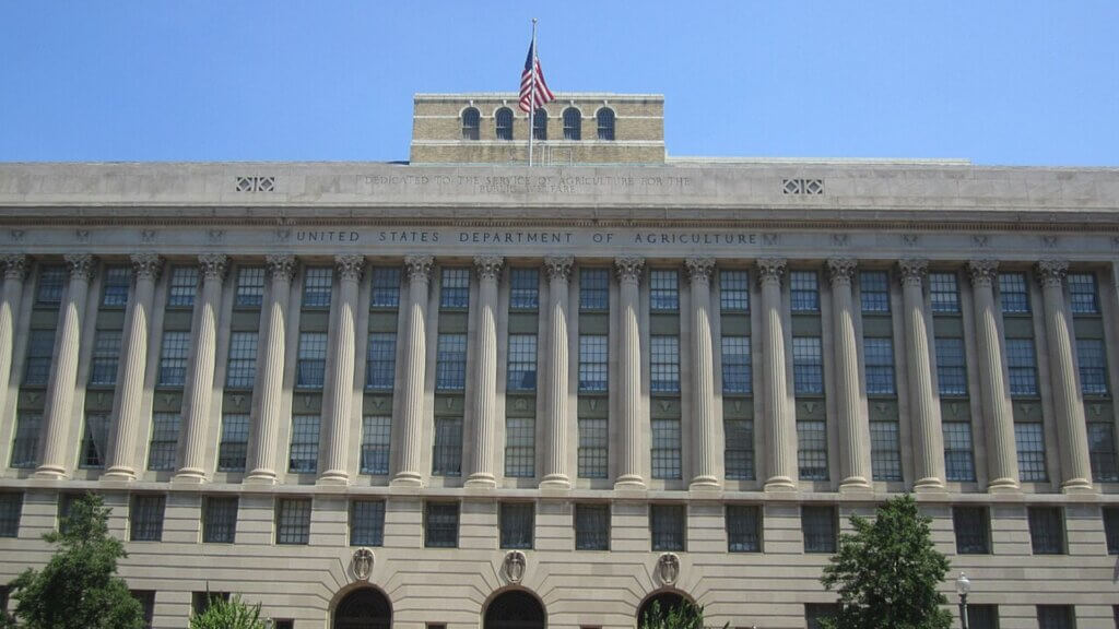The front facade of the United States Department of Agriculture building features tall white columns and large windows.