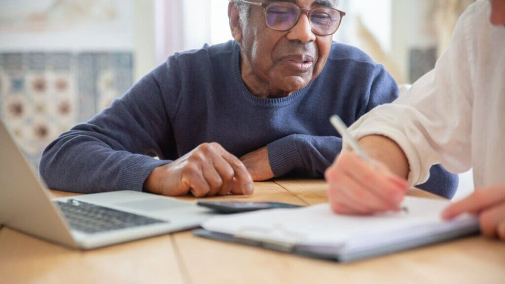 Older man reviews paperwork at a table while another person takes notes, with a laptop nearby