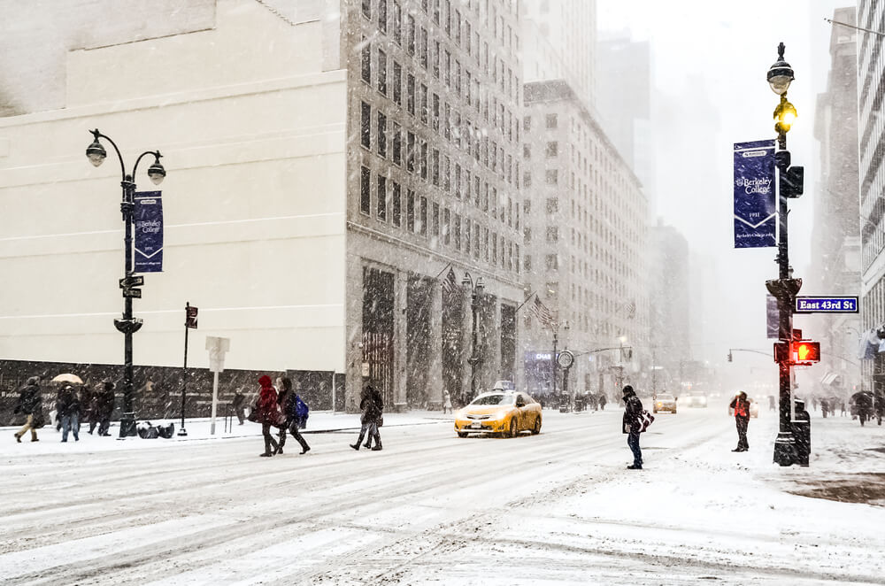 Pedestrians cross a snow-covered Manhattan street as a yellow taxi drives through heavy snowfall.