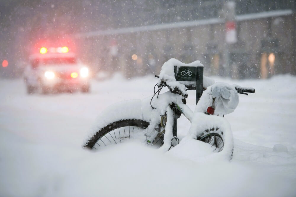 Snow-covered bicycle parked on a New York City street during a blizzard as emergency lights glow in the background.
