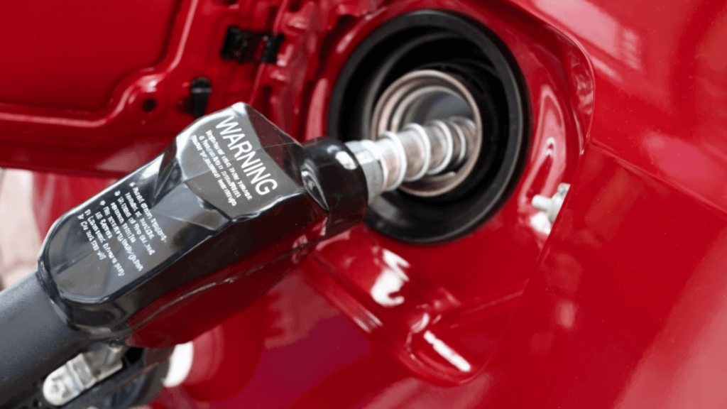 Close-up of a black fuel nozzle inserted into the gas tank of a red car, with a white warning label visible on the handle.