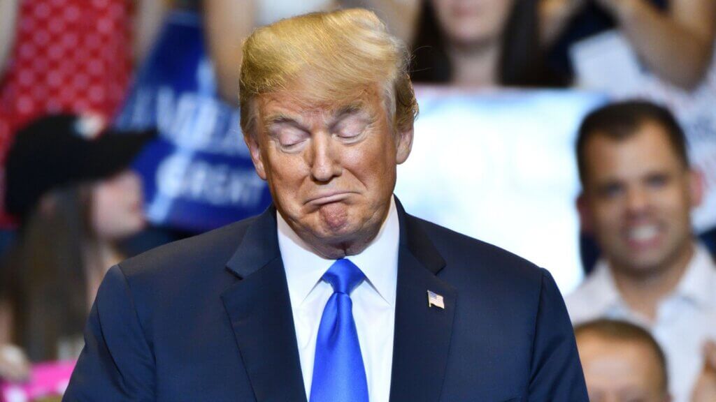 Donald Trump with eyes closed and pursed lips while standing before a crowd at a campaign rally.