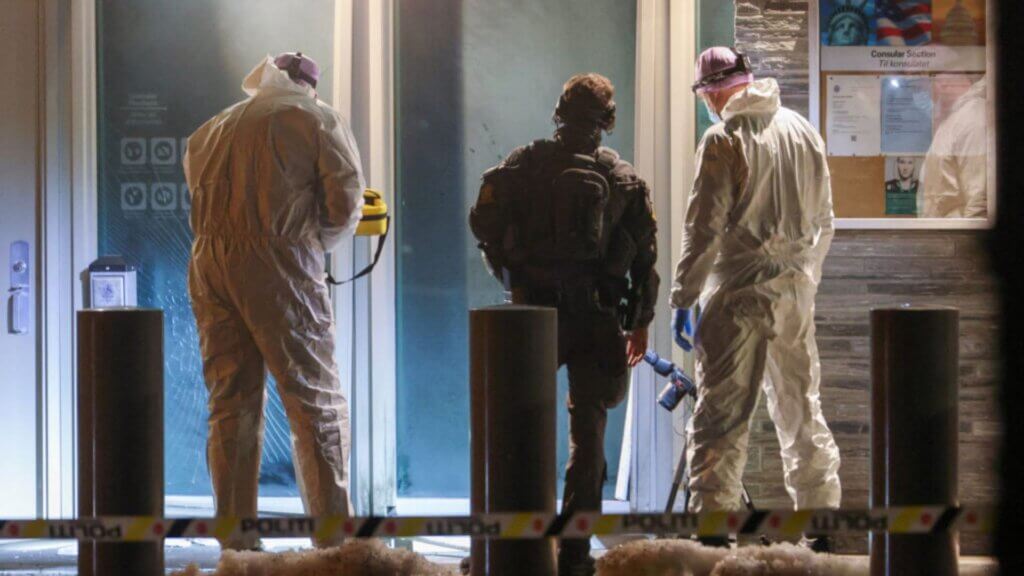 Forensic investigators and a police officer examine a damaged entrance outside a U.S. consular building at night.