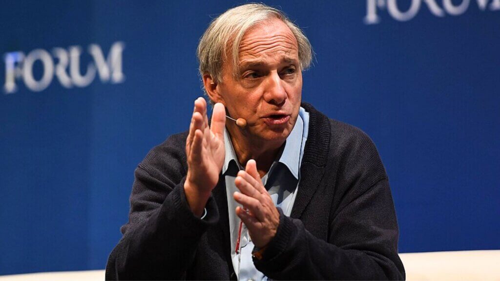 Ray Dalio gesturing during a forum discussion on stage against a blue backdrop.