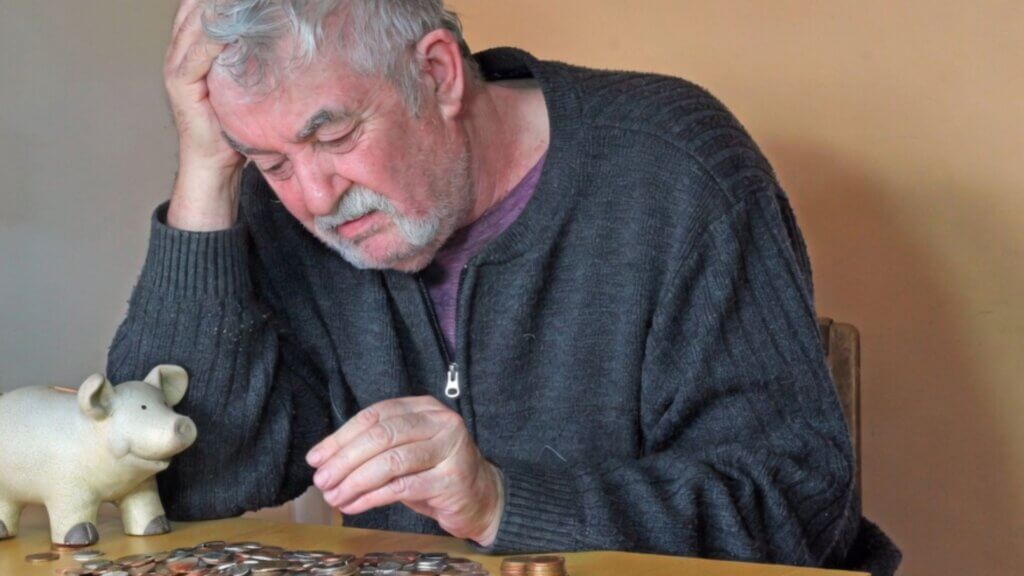 Elderly man counting coins beside a piggy bank with his head in his hand.