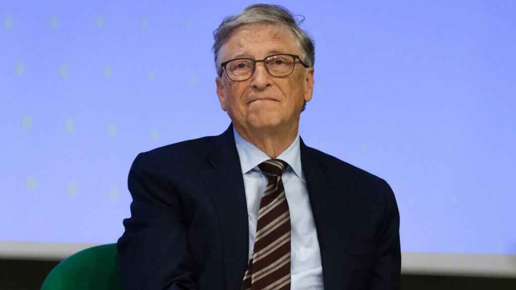 Bill Gates seated on stage during a panel discussion, wearing a suit and tie.