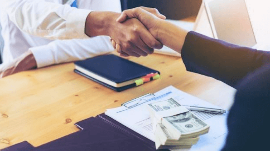 Close-up of two people shaking hands over a wooden table with a large stack of cash and paperwork visible.