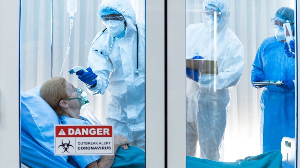Medical staff in full protective suits monitoring an elderly patient with oxygen mask in an isolation hospital ward.