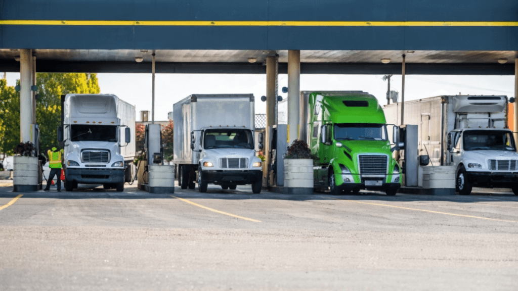 A row of several large semi-trucks, including white and bright green models, parked under the awning of a commercial truck stop.
