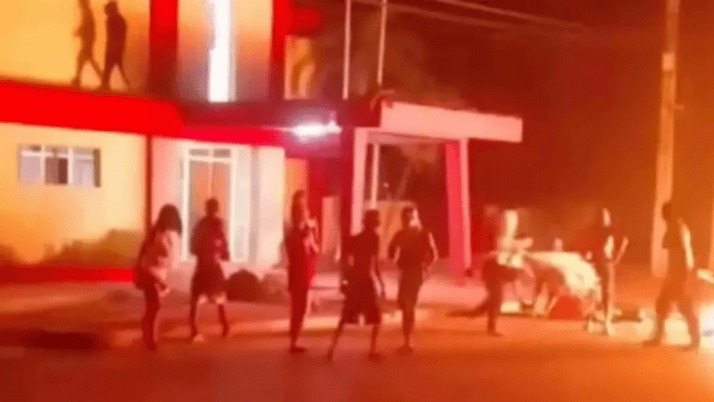 A nighttime photo of several individuals standing in front of a building that is glowing with intense red and orange light from a fire inside.