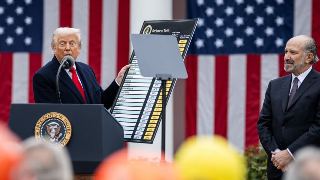 Donald Trump speaking at podium while holding tariff chart with U.S. flags behind him.