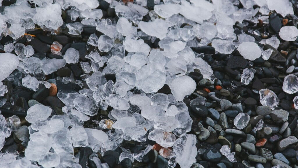 Large chunks of ice scattered across dark pebbles on the ground after a hailstorm.