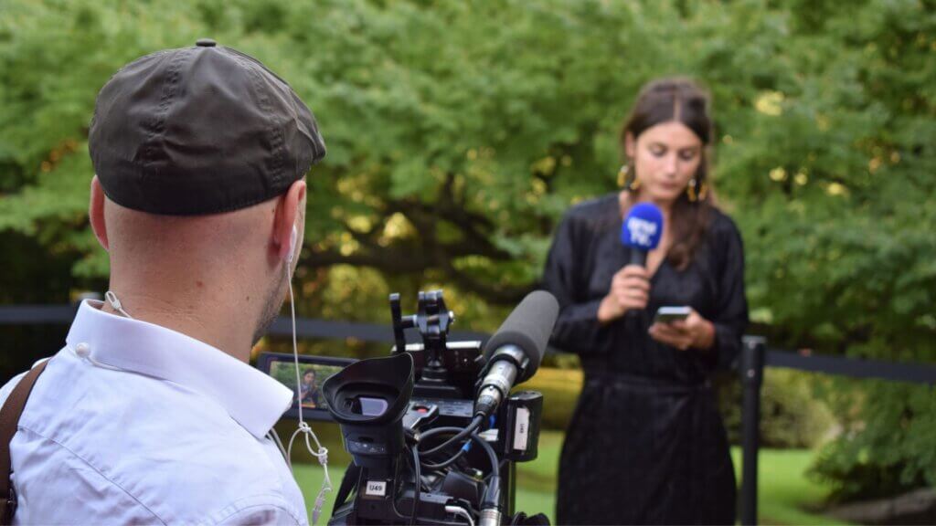 Television reporter holding microphone while cameraman films her outdoors in a park setting.