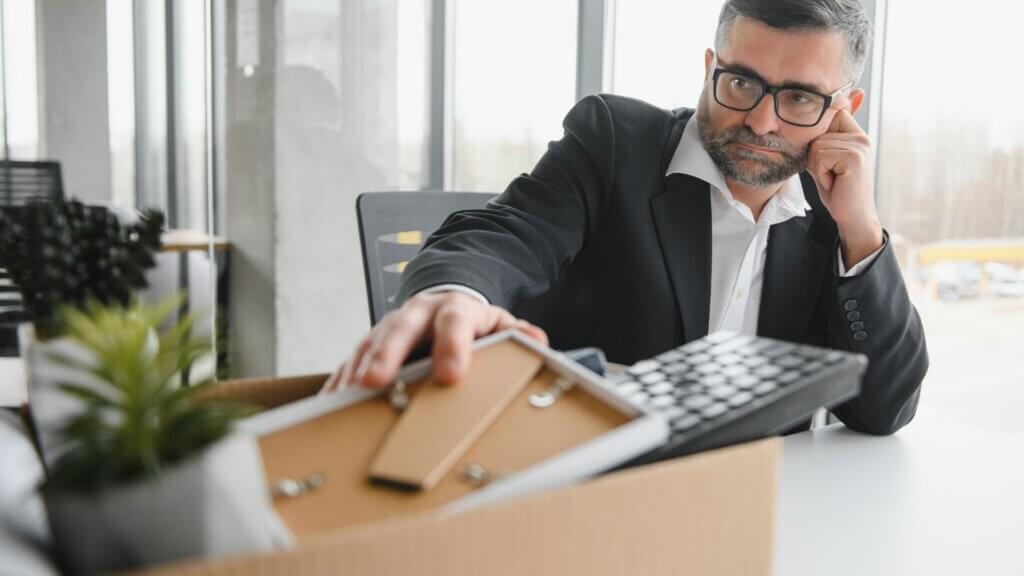 Older male office employee placing personal belongings and a calculator into a box after losing his job