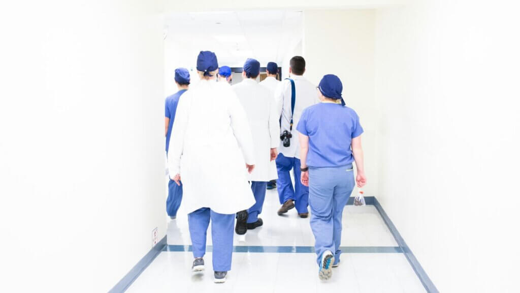 Group of medical staff in scrubs and white coats walking down a hospital corridor.