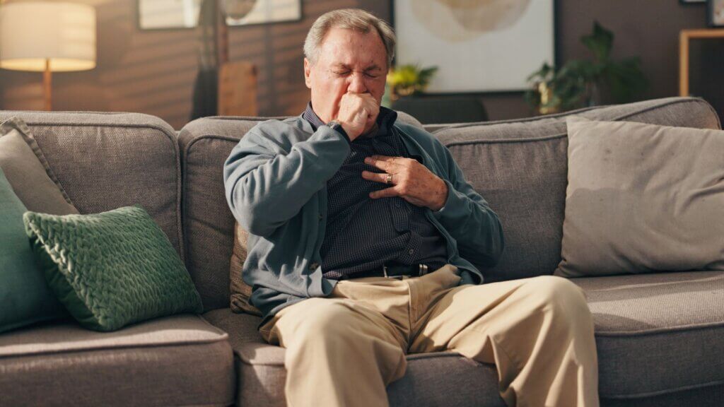 Older man sitting on a couch coughing into his hand while holding his chest at home.