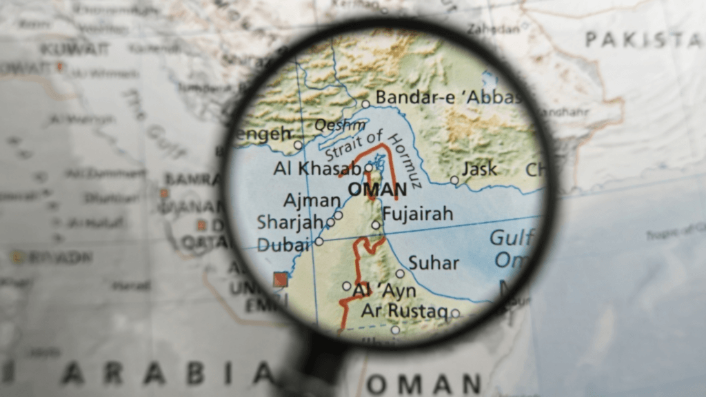 A magnifying glass held over a world map, focusing on Oman and the strategic shipping route of the Strait of Hormuz.