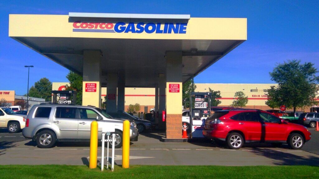 Cars lined up at pumps under a Costco Gasoline station canopy at a warehouse store parking lot.