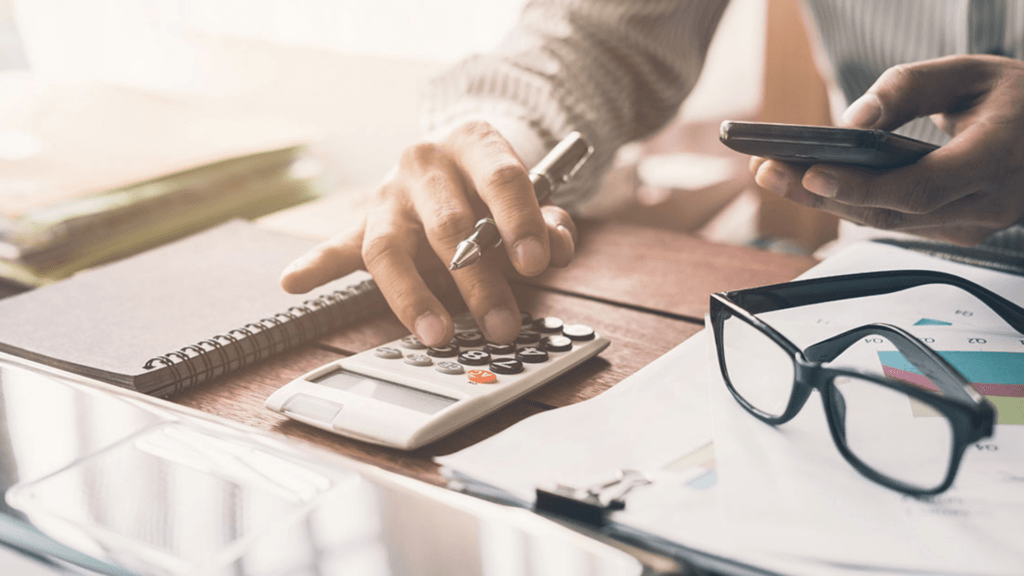 A person using a calculator and a smartphone at a desk covered in financial documents, charts, and a notebook to calculate taxes.