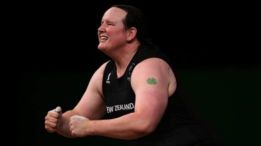 Weightlifter Laurel Hubbard in a black singlet, exhaling with effort and showing a green four-leaf clover tattoo on her upper arm during a competition.