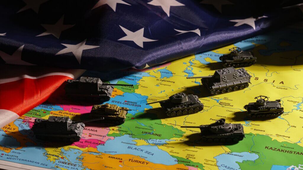 Toy tanks on a map of Europe beneath a US flag, illustrating geopolitical conflict.