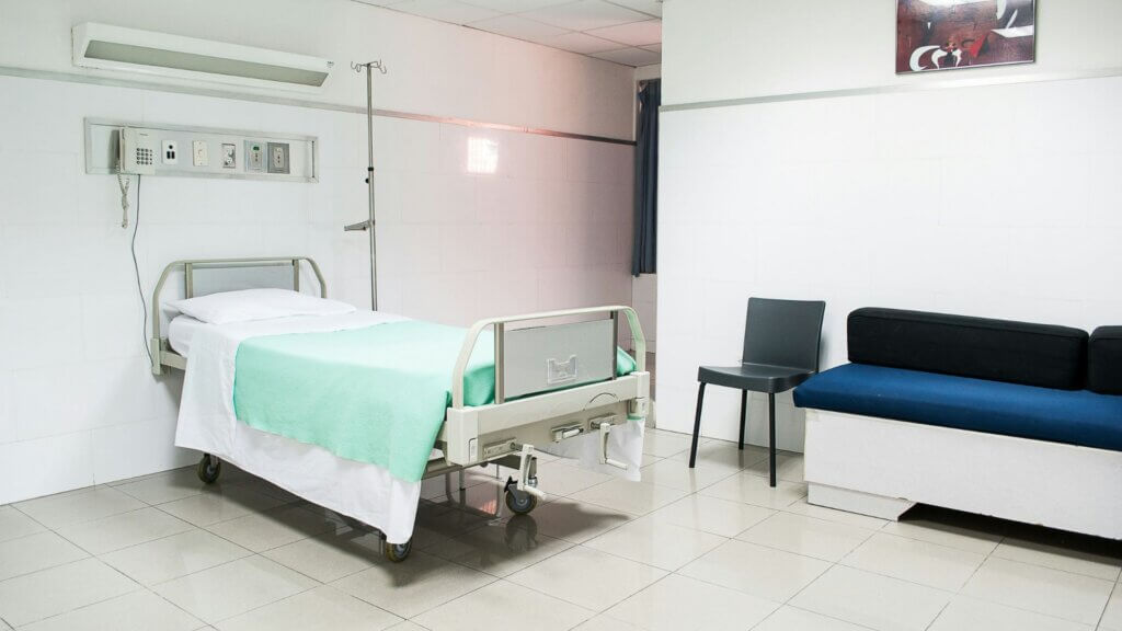 Empty hospital room with a bed, IV stand, and seating area near the wall.