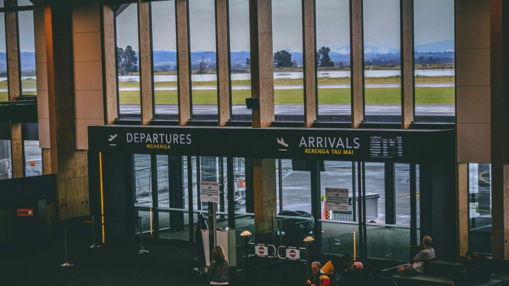 Travelers walking through a busy airport terminal with large windows.