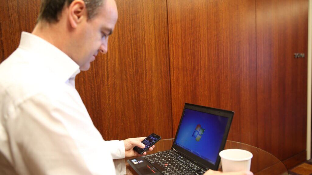 Man using a smartphone next to an open laptop on a table.
