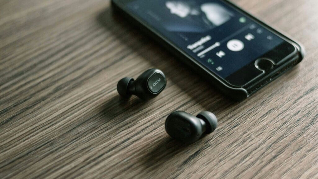 Wireless earbuds placed next to a smartphone playing audio on a wooden table.