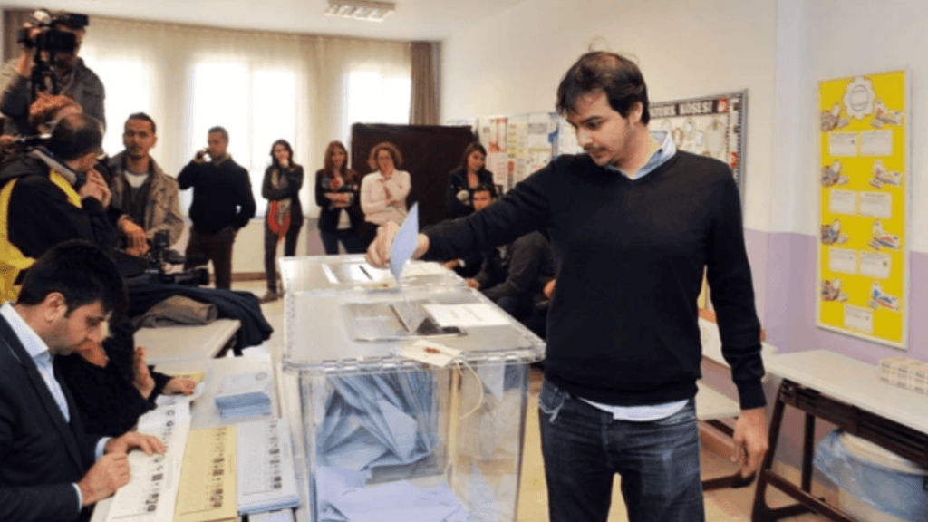 A man casting a paper ballot into a clear voting box at a polling station with observers in the background.