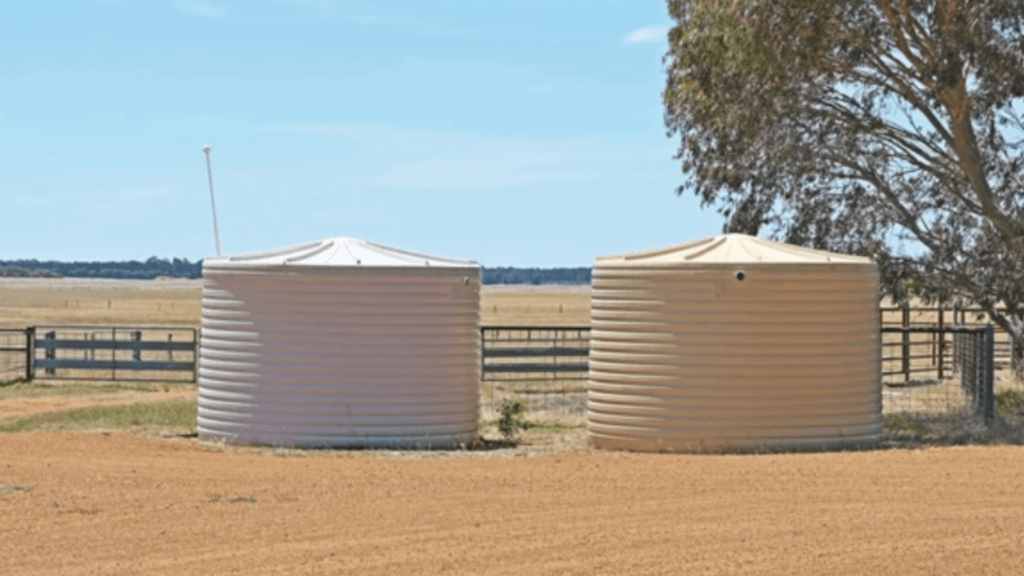 Two large plastic water storage tanks sitting on a rural property under a clear blue sky.