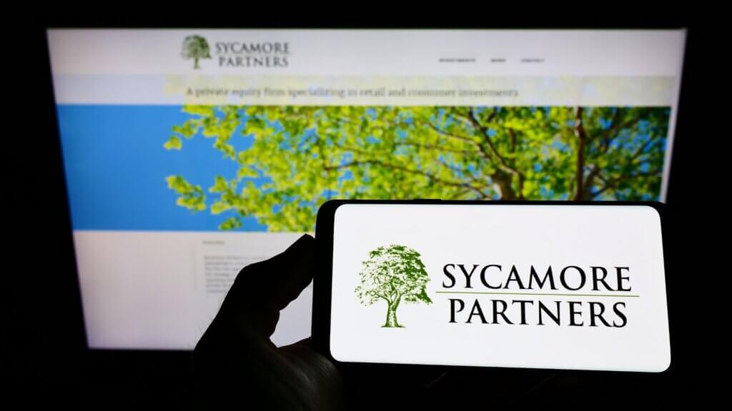 Smartphone displaying the Sycamore Partners logo held in front of the company website on a computer screen