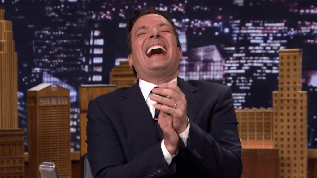 A close-up of Jimmy Fallon laughing heartily and clapping his hands while sitting at his desk.