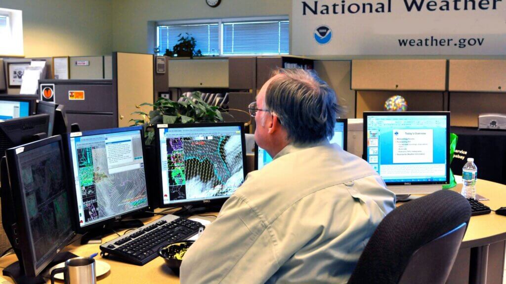 Meteorologist monitoring weather radar maps on multiple computer screens in forecast office.