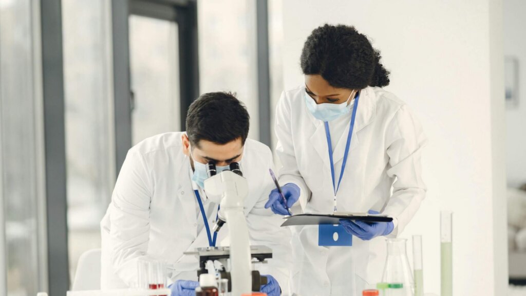 Scientist looking through a microscope while a colleague assists in a laboratory.