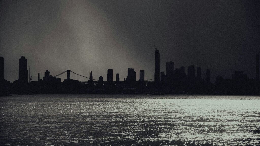 Dark city skyline across water under stormy sky.
