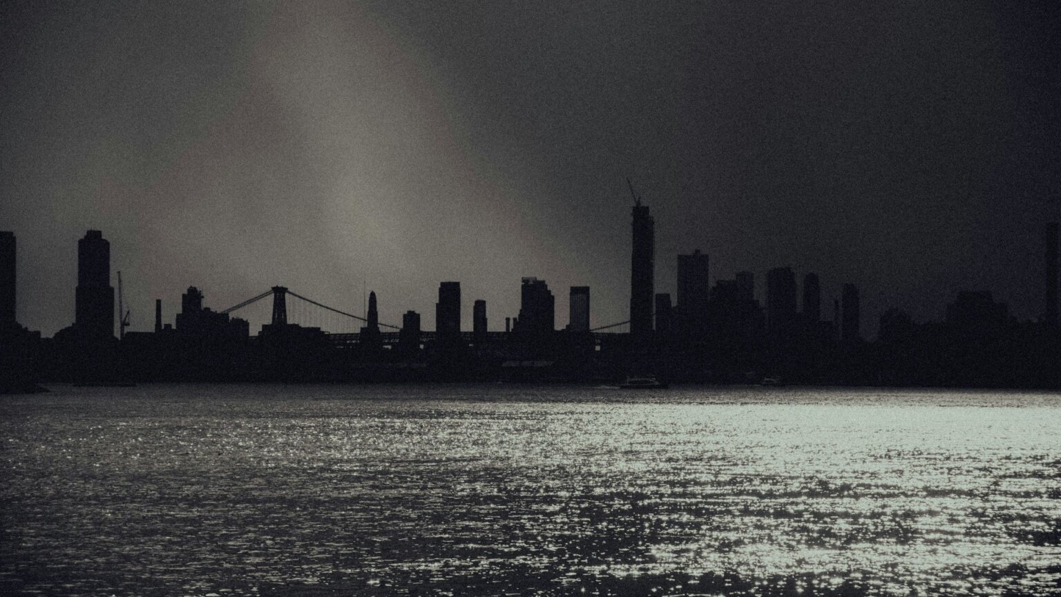 Dark city skyline across water under stormy sky.