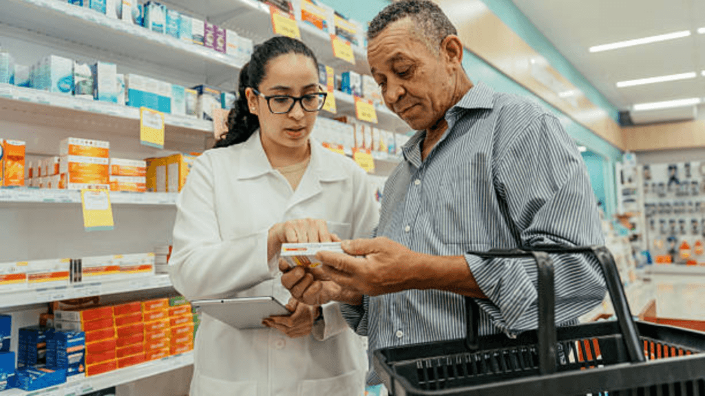 A pharmacist in a white coat using a tablet to explain medication details to a senior man in a pharmacy aisle.