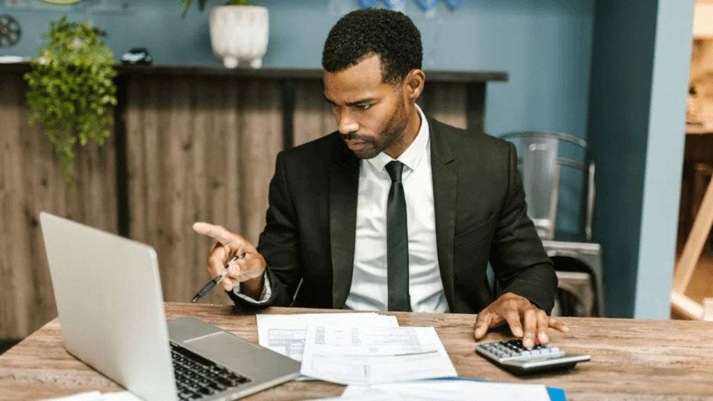 A man in a dark suit works on his laptop with papers and a calculator.