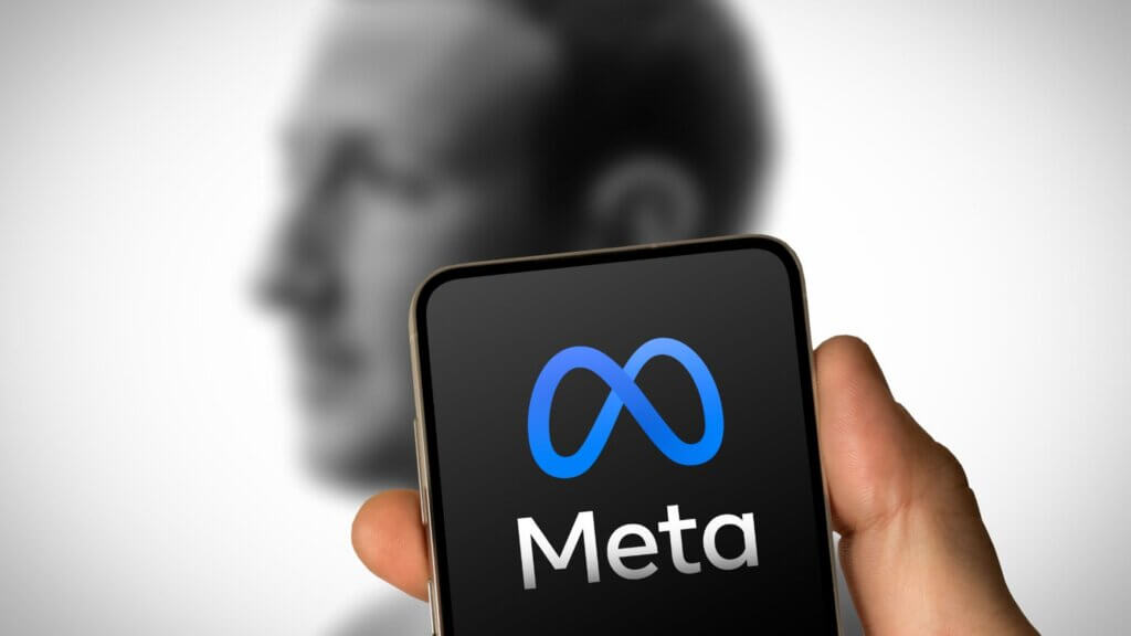 A cellphone showing meta´s logo and mark zuckerberg in the back.
