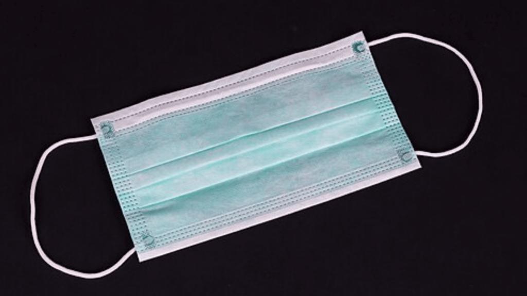 A green disposable surgical mask with white ear loops lies flat against a solid black background.