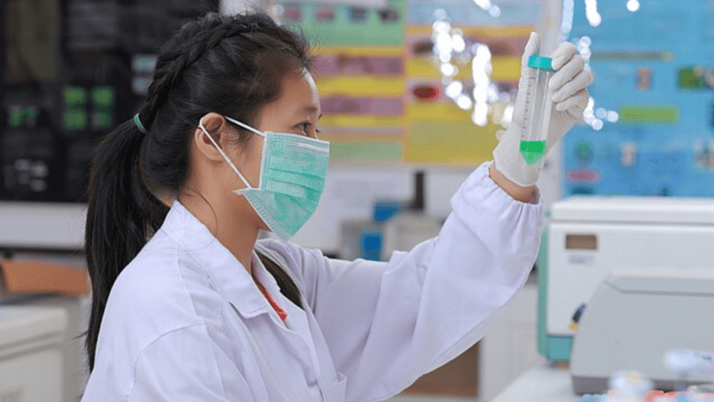 A lab technician wearing a white coat, green surgical mask, and gloves holds up a test tube containing a green liquid in a laboratory setting.