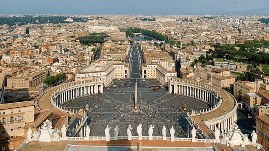 Wikimedia An aerial view of St. Peter’s Square in Vatican City, showing the large circular colonnades, central obelisk, and the surrounding city of Rome in the background.