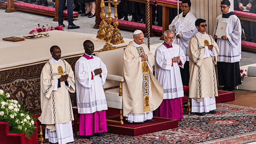 Group of Catholic clergy dressed in cream and white liturgical robes standing on a raised platform during a religious ceremony, with an ornate chair and decorative element.
