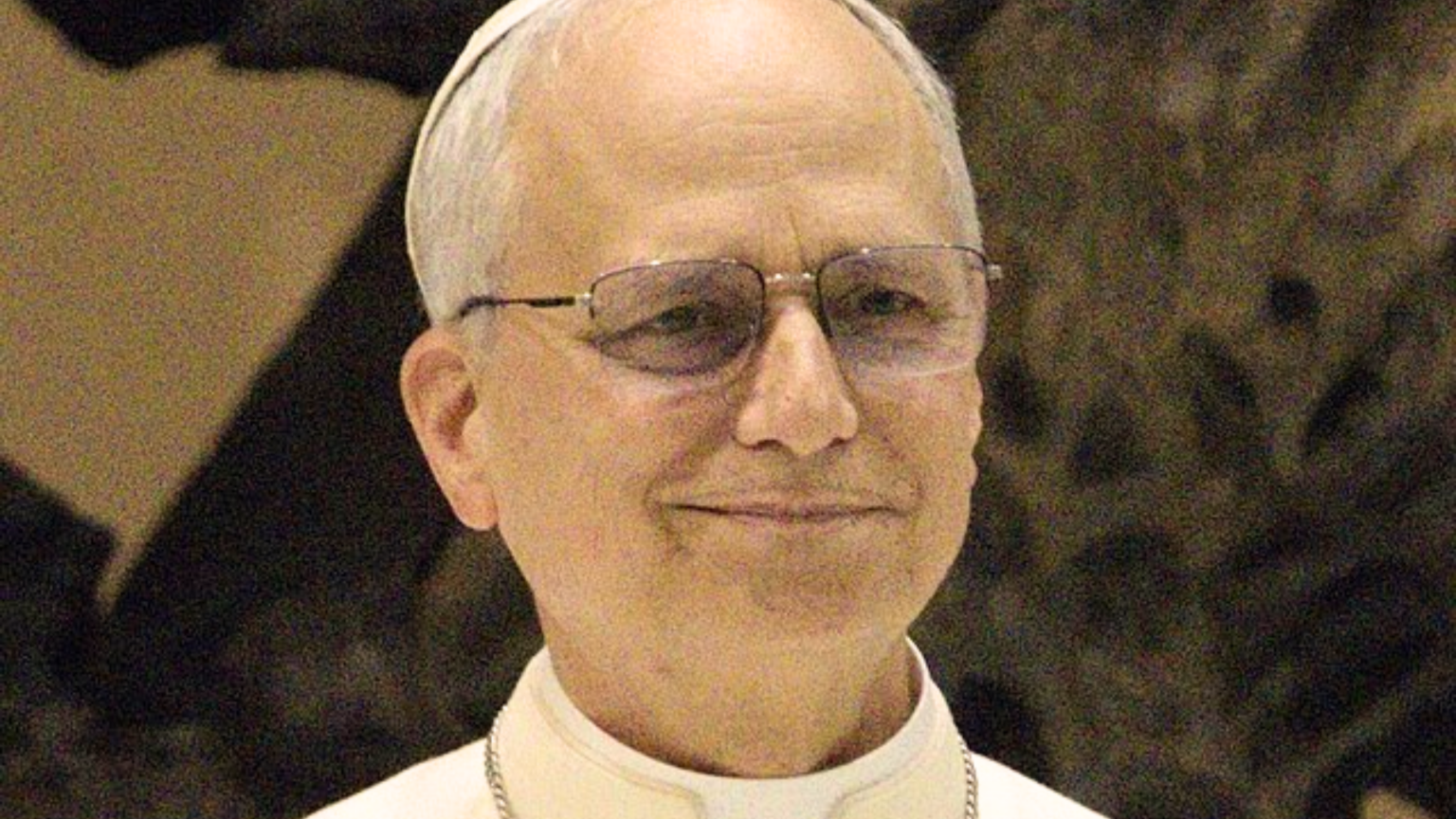 Pope Leo XIV wearing white papal robes and a skullcap and glasses, smiling gently.