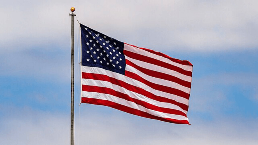 The American flag waving from a flagpole against a backdrop of soft, blue-and-white clouds.