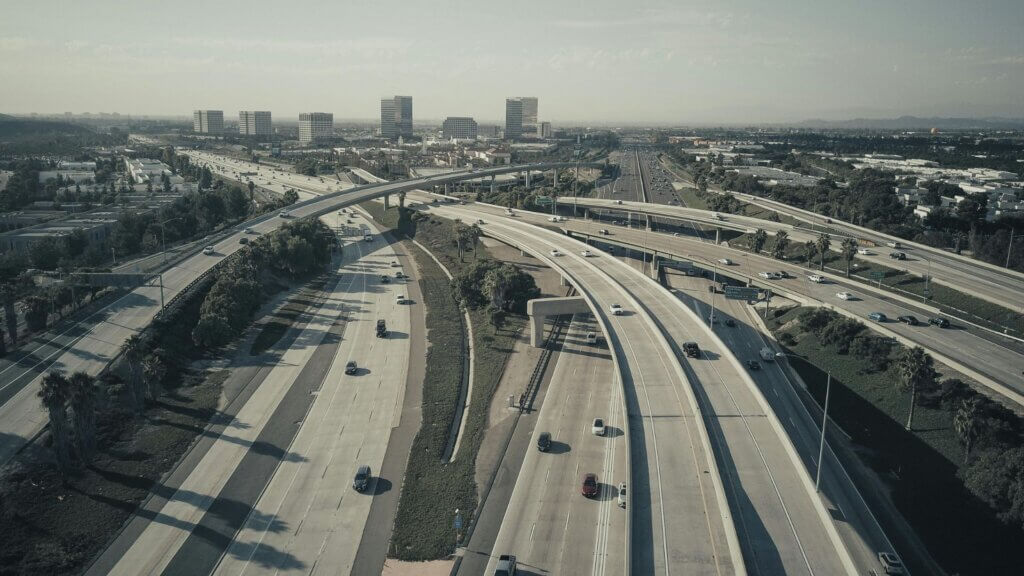 Aerial view of a sprawling freeway interchange with multiple overpasses, surrounded by green spaces and distant city buildings.