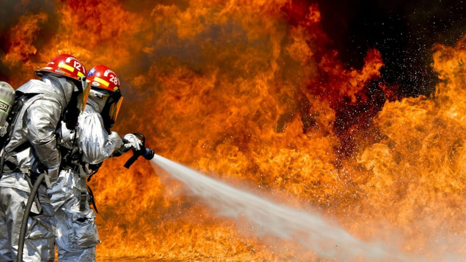 A photo of two firemen wearing protective gear and killing a huge fire.