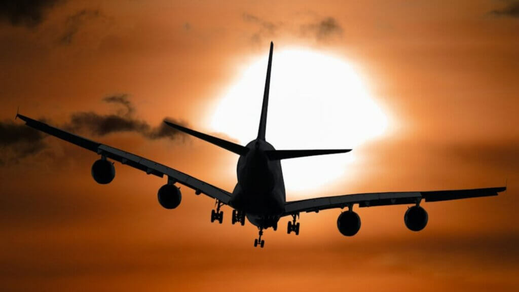 Silhouette of an airplane flying towards a large setting sun, with an orange and brown sky.