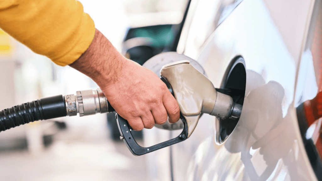 A close-up of a hand holding a fuel nozzle while refueling a silver car at a gas station.